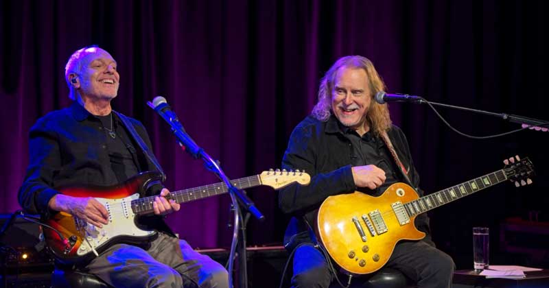 Two musicians playing guitars on stage, smiling.